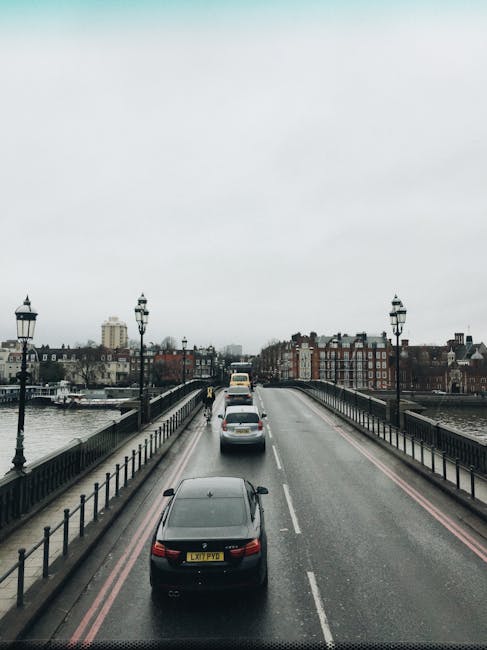 Aerial view of a city bridge with a grey overcast sky, showing a black car in the foreground and several other vehicles, including a van, moving along the road. The bridge features black iron railings on both sides and decorative street lamps evenly spaced along its length. On either side of the river, there are residential and commercial buildings with various architectural styles, some with brick facades and others with modern designs. The scene illustrates typical urban traffic flow, relevant to house removals and furniture transport services such as those provided by [COMPANY_NAME]. The image captures the urban environment where professional removals may operate during home relocation projects, supporting logistics for moving furniture and boxes across city areas, including those with tight access or limited space, as discussed in the Battersea Park house removals guide for tight access, BATTERSEA.