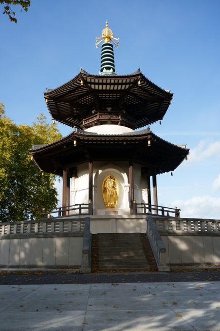 A traditional multi-tiered pagoda-style structure with dark wooden eaves and decorative gold accents, situated on a raised platform accessed by a short staircase. The building is enclosed by a curved stone balustrade, and the surrounding area features paving stones and some trees with autumn foliage. The sky is clear with a few clouds. This scene depicts a heritage or religious site, with no visible furniture or moving activity. The image emphasizes the architecture and setting, which could be relevant for home relocation or cultural moves within the context of removals services.