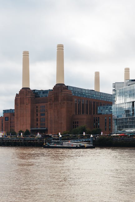 Aerial view of a city bridge with a grey overcast sky, showing a black car in the foreground and several other vehicles, including a van, moving along the road. The bridge features black iron railings on both sides and decorative street lamps evenly spaced along its length. On either side of the river, there are residential and commercial buildings with various architectural styles, some with brick facades and others with modern designs. The scene illustrates typical urban traffic flow, relevant to house removals and furniture transport services such as those provided by [COMPANY_NAME]. The image captures the urban environment where professional removals may operate during home relocation projects, supporting logistics for moving furniture and boxes across city areas, including those with tight access or limited space, as discussed in the Battersea Park house removals guide for tight access, BATTERSEA.