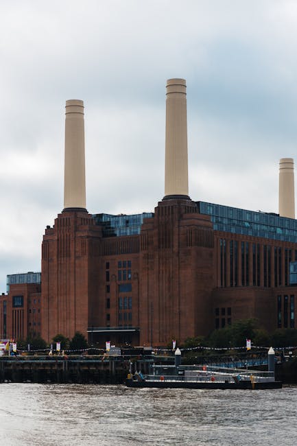 A view of a large industrial-style building with three tall chimney stacks situated along the riverbank, adjacent to a modern glass office building, under an overcast sky. In the foreground, the river